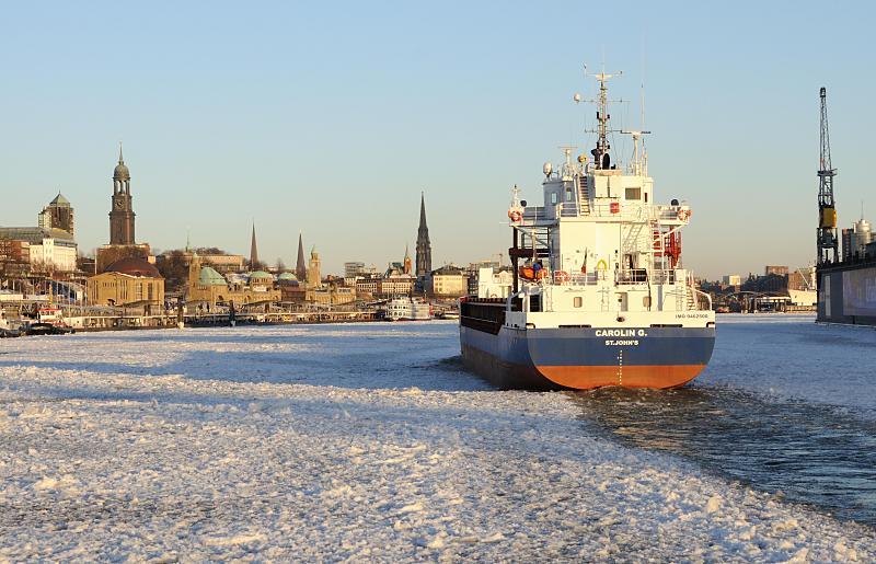 Fotos vom Hamburger Winter : Eisgang auf der Elbe : Hamburger Bilder Hamburg Bilder vom Winter im Hamburger Hafen - Frachter auf der Elbe, Eisgang - Hamburger Kirchtrme. 011_78_5837 Das 88 m lange und 12 m  breite Frachtschiff CAROLIN G. luft in den Hamburger Hafen ein. Die Elbe ist dicht mit Treibeis bedeckt. Die Abendsonnen scheint auf das Panorama mit den Trmen der Hansestadt Hamburg - ganz links der Kirchturm vom Hamburger Wahrzeichen die St. Michaeliskirche, auch Michel genannt; daneben die Spitze vom Rathausturm, die St. Petri-Kirche, die Jakobikirche, der Uhr- und Pegelturm der St. Pauli Landungsbrcken, die Gustav-Adolf-Kirche und im Bildzentrum der Turm der St. Nikolaikirche. 