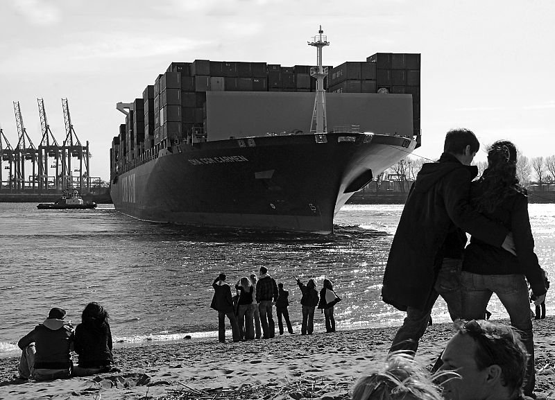 11_17453 das Containerschiff CMA CGM Carmen wird  hoch beladen von zwei Schleppern in den Hamburger Hafen gebracht - der Containerriese hat eine Lnge von ca. 300m und eine Breite von 40m; der Frachterf kann 6627 TEU / 20 Fuss Container als Ladung an Bord nehmen. Am Elbstrand stehen und sitezen Hamburger, die das Schiffsmanver interessiert beobachten.  www.christoph-bellin.de