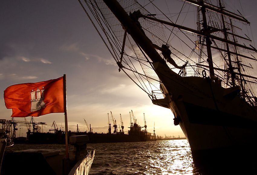 011_15708 Sonnenuntergang im Hamburger Hafen; die Hamburg Fahne strahtl in der Abend Sonne - vom Grosssegler am Kai der Landungsbrcken sind Bug und Masten in der Silhouette zu erkennen - im Hintergrund Krne der Werft an der Elbe.  www.fotograf-hamburg.de