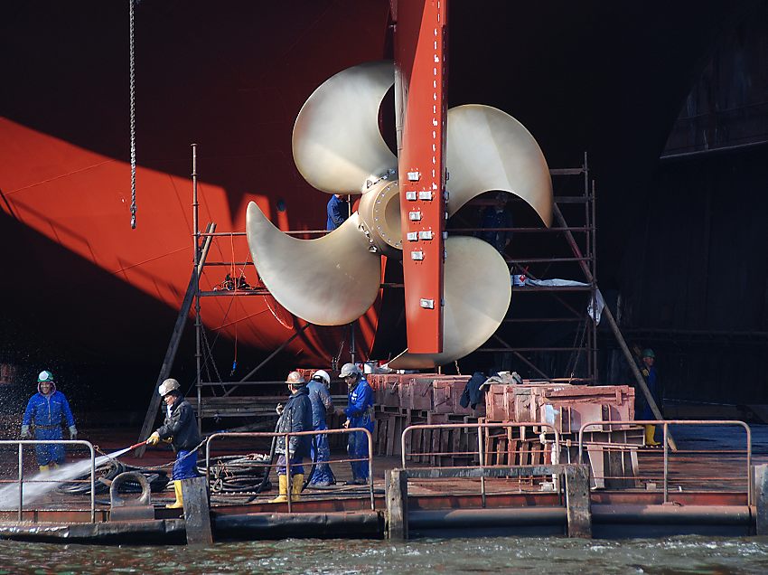 011_22118/00 im Trockendock der Schiffswerft Blohm + Voss bekommt ein Frachtschiff eine neue Schiffsschraube aus glnzendem Messing - Hafenarbeiter reinigen die Werftanlage  mit einem Wasserstrahl. www.fotograf-hamburg.de