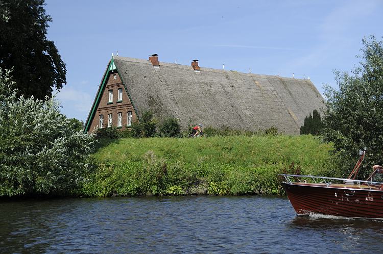 011_26063 Ein strohgedecktes Bauernhaus hinter dem Elbdeich der Dove-Elbe; das Strohdach des Hauses ragt hinter dem Deich hervor auf dem ein Radrennfahrer mit seinem Fahrrad fhrt. -auf dem Wasser der Dove-Elbe kommt ein holzgeklinkertes Motorboot, dessen Bug und Reling schon zu erkennnen ist.     www.christoph-bellin.de