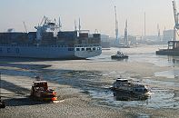 011_26072 Hamburger Winter - die Elbe ist mit Eisschollen bedeckt; eine Hafenfhre und ein Schiff der Hamburger Hafenrundfahrt bahnen sich ihren Weg durch den Eisgang. Ein voll beladener Containerriese wird mit dem Heck von einem Schlepper in das Hafenbecken TOLLERORT gezogen. Im Hintergrund Hafenkrne an den Kaianlagen des Hamburger Hafens. www.hamburg-fotograf.com