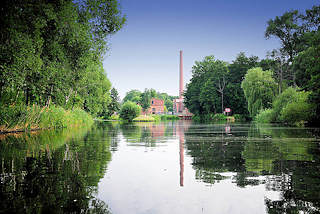 0436 Alte Fabrikgebäude mit hohem Ziegelschornstein von Neu Kaliß spiegeln sich im Wasser des Elde Müritz Wasserwegs. Hohe Bäume und Schilf am Ufer des Wasserwegs.