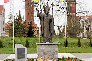 0113 Statue Papst Johannes Paul II. in Pieniężno, Polen. Im Hintergrund die Kirchtürme der evangelischen und katholischen Kirche und der restaurierte Kupferturm vom Rathaus.