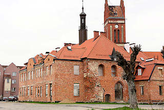 0114 Altes Rathaus von Mehlsack / Pieniężno, Polen. Das Backsteingebäude steht leer, die Fenster sind vernagelt - Kirchturm der katholischen Kirche der Stadt.