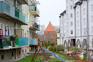 0230 Mehrstöckige Wohnhäuser mit farbigen Balkons - Wiese mit Kinderspielplatz; im Hintergrund der sogen. Storchenturm, Teil der Befestigungsanlage von Dobre Miasto / Guttstadt, Polen.